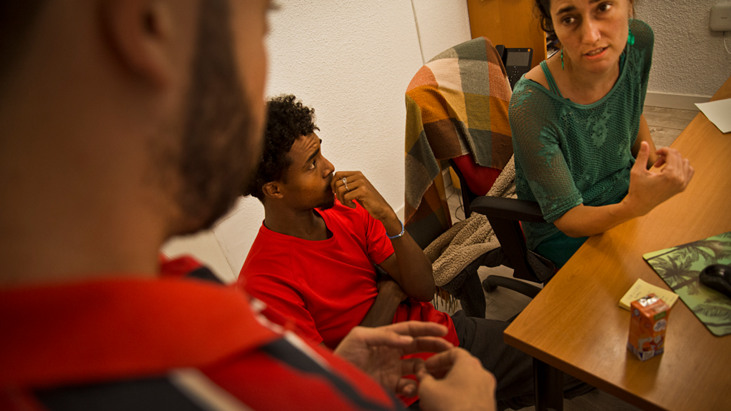 Una orientadora sentada en su escritorio, frente el ordenador, junto a dos jóvenes extutelados racializados. Uno de ellos está sentado a su lado y el otro, de pie, como si le trasladara alguna duda puntual.
