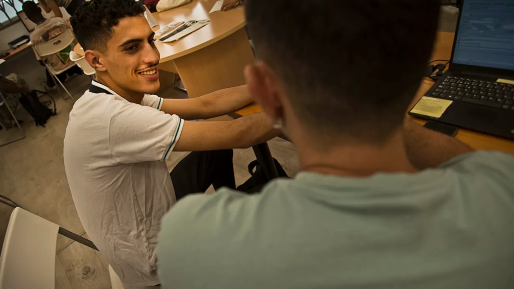 En un aula de Fundación ISOS, un joven extutelado racializado, de cara, observa a su educador social. Están sentidos en una mesa ante un ordenador.